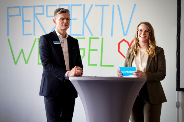 Rückblick auf das pädia Pädiatrie-Event: Zwei Moderatoren stehen an einem Stehtisch vor der "Perspektivwechsel"-Schriftwand. Die Frau hält eine PädiaAkademie-Mappe, der Mann trägt ein Namensschild und ein Ansteckmikrofon.