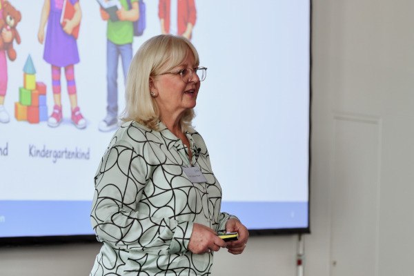 Rückblick auf das pädia Pädiatrie-Event: Eine Referentin hält einen Vortrag vor einer Leinwand mit kindlichen Illustrationen verschiedener Altersgruppen, darunter die Beschriftungen "Kleinkind" und "Kindergartenkind".