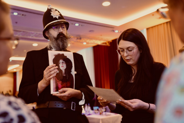 Rückblick auf das pädia Pädiatrie-Event: Ein als britischer Bobby verkleideter Spielleiter der Sherlock Holmes Academy hält ein Foto einer Verdächtigen in die Höhe. Daneben liest eine Teilnehmerin konzentriert einen Hinweiszettel. Im Hintergrund ist der festlich beleuchtete Veranstaltungssaal zu erkennen.