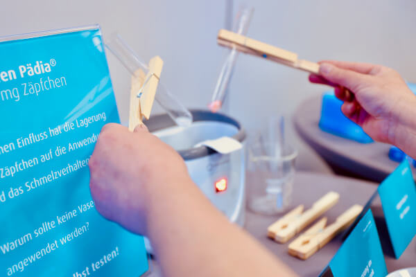 Rückblick auf das pädia Pädiatrie-Event: Zwei Teilnehmerinnen experimentieren an einer PädiaLab-Station mit Ibuprofen Pädia Zäpfchen. Mit Holzwäscheklammern und Reagenzgläsern untersuchen sie das Schmelzverhalten und die Lagerung. Daneben steht ein blaues Informationsschild mit Fragen wie "Welchen Einfluss hat die Lagerung der Zäpfchen?" und "Warum sollte keine Vaseline angewendet werden?"