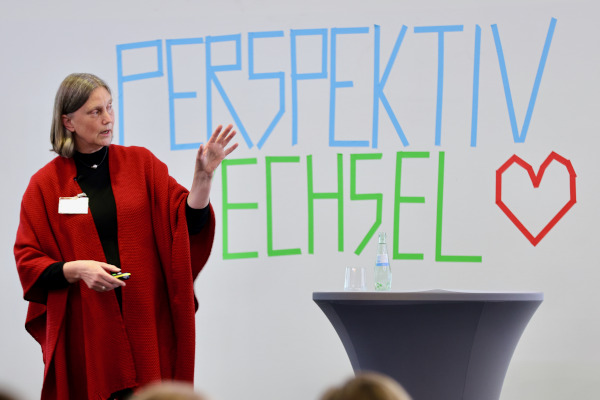 Rückblick auf das pädia Pädiatrie-Event: Dr. Vetye hält den Abschlussvortrag vor der "Perspektivwechsel"-Schriftwand mit dem roten Herz. Sie gestikuliert lebhaft und hält einen Präsentationsklicker in der Hand. Ihr Vortrag handelt von ihrer Arbeit bei Apotheker ohne Grenzen in den benachteiligten Stadtteilen von Buenos Aires.